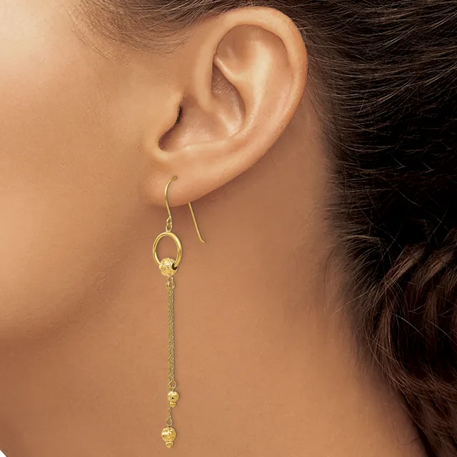 A woman wearing gold dangle earrings with small spherical beads on thin chains, shown from the side profile near her ear.
