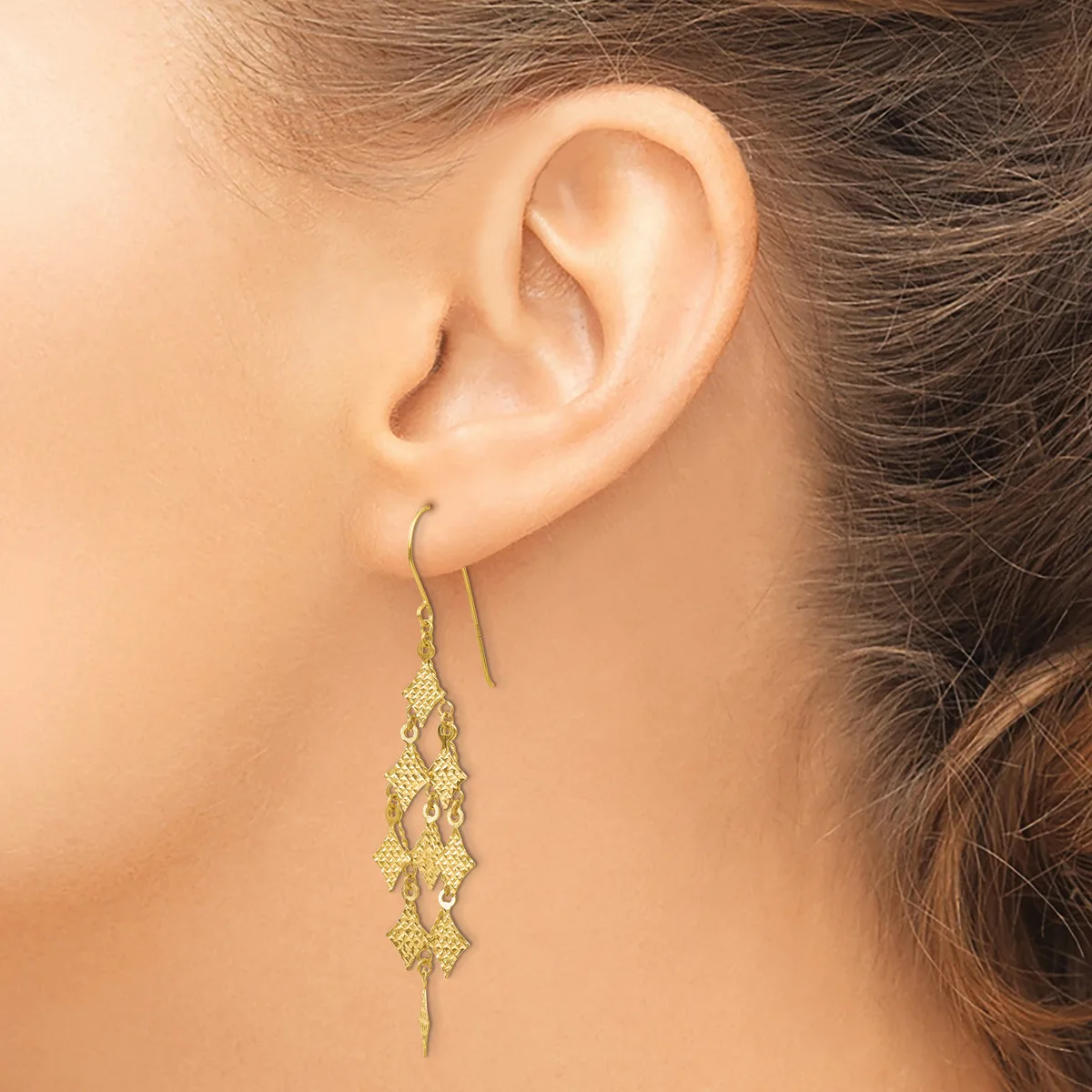 A person wearing gold-toned drop earrings with textured diamond-shaped charms hanging from curved wire hooks.