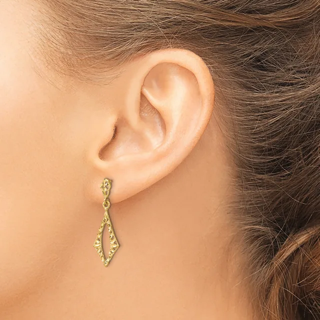 A woman wears a pair of gold-toned drop earrings with elongated diamond shapes and textured surfaces, displayed on her pierced earlobes.
