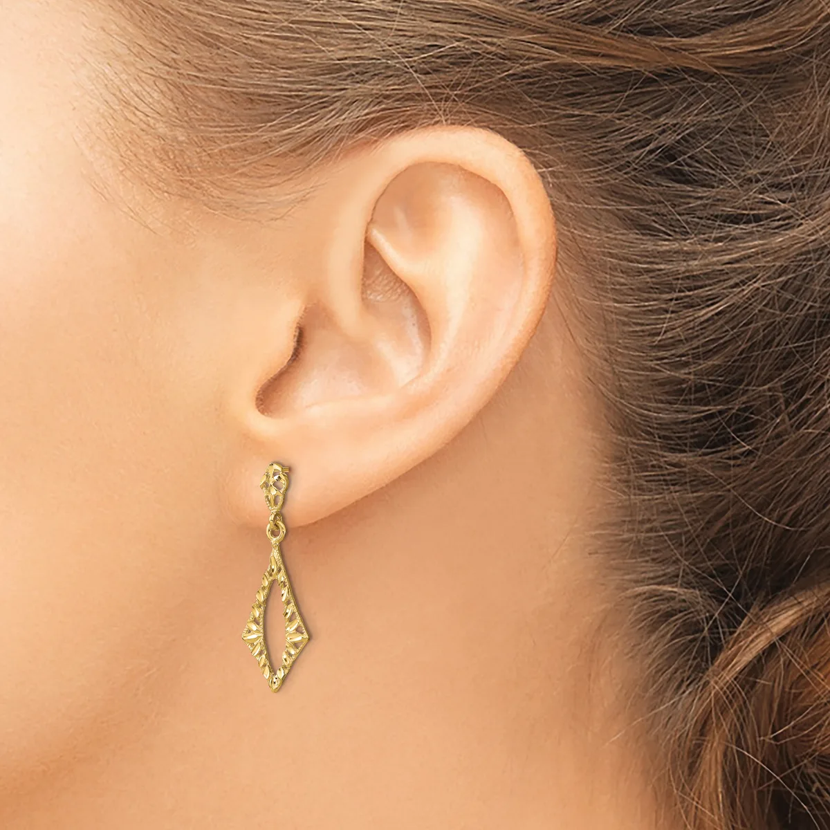 A woman wears a pair of gold-toned drop earrings with elongated diamond shapes and textured surfaces, displayed on her pierced earlobes.