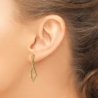 A woman wears a pair of gold-toned drop earrings with elongated diamond shapes and textured surfaces, displayed on her pierced earlobes.