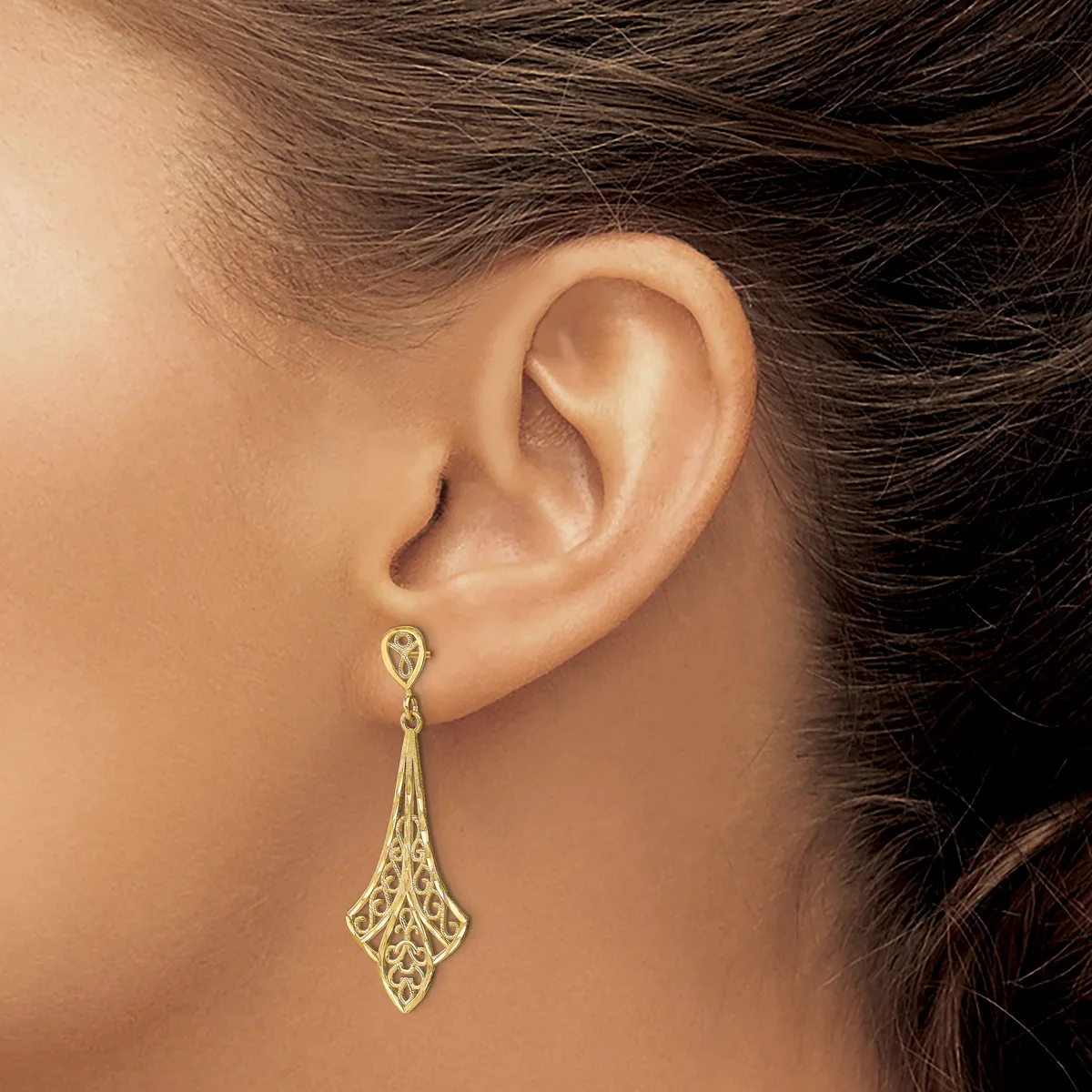 A woman wearing a gold filigree teardrop-shaped dangle earring with intricate openwork detailing.