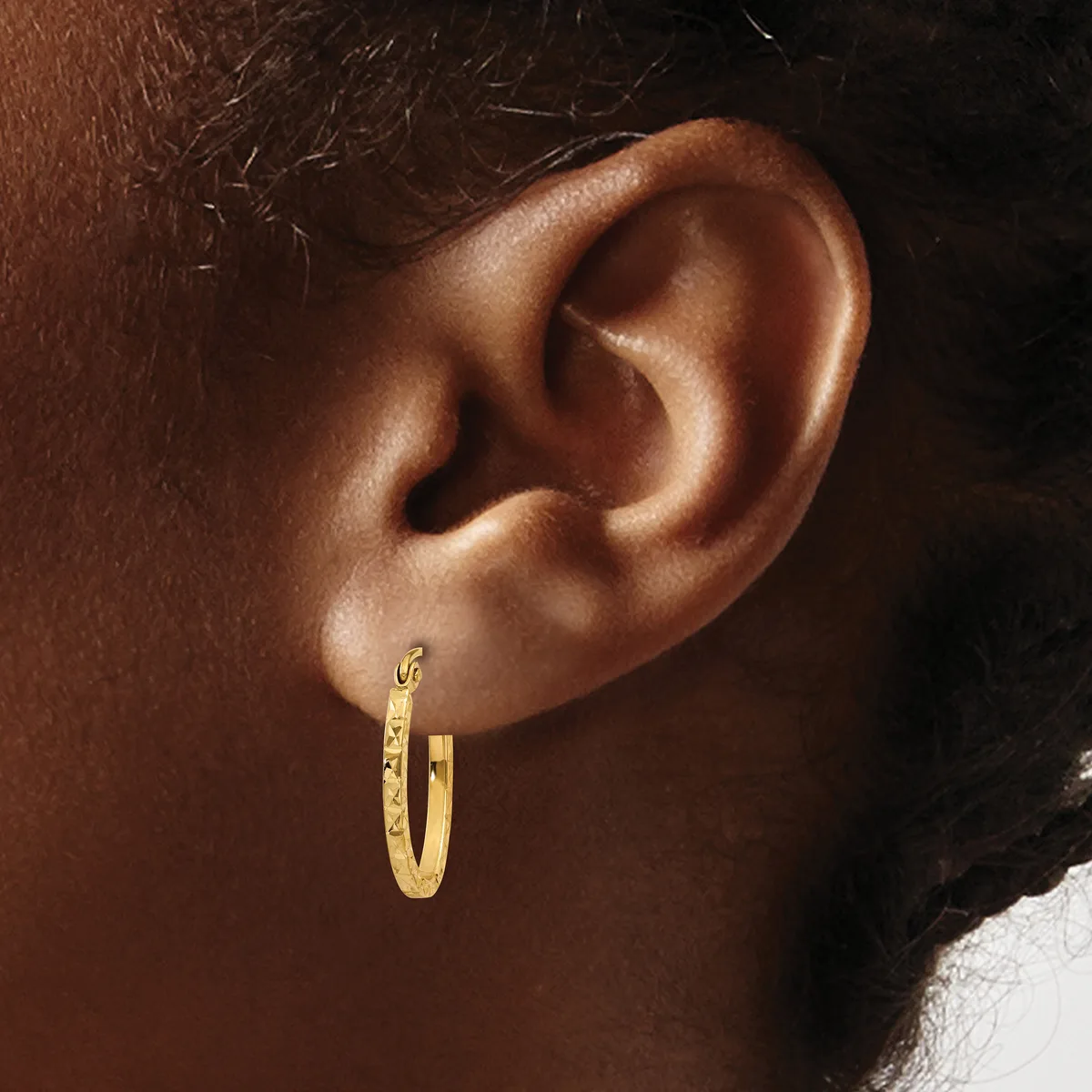A close-up side view of a person's ear wearing a textured gold hoop earring with a small hallmark stamp visible on the post.