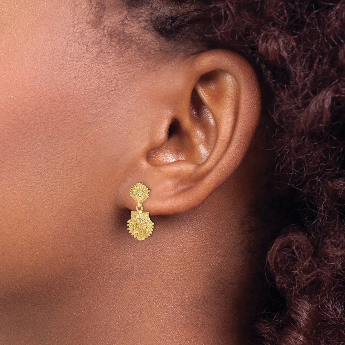 A person wearing gold-textured drop earrings with a scalloped shell-like design on both the upper and lower portions of each earring.