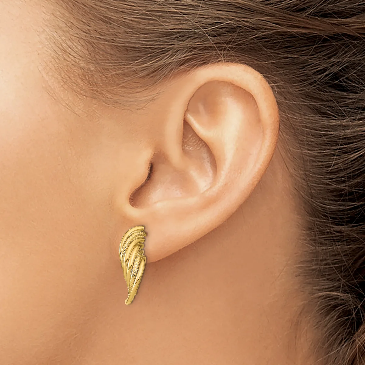 A gold wing-shaped earring with textured feather detailing hangs from a pierced earlobe.