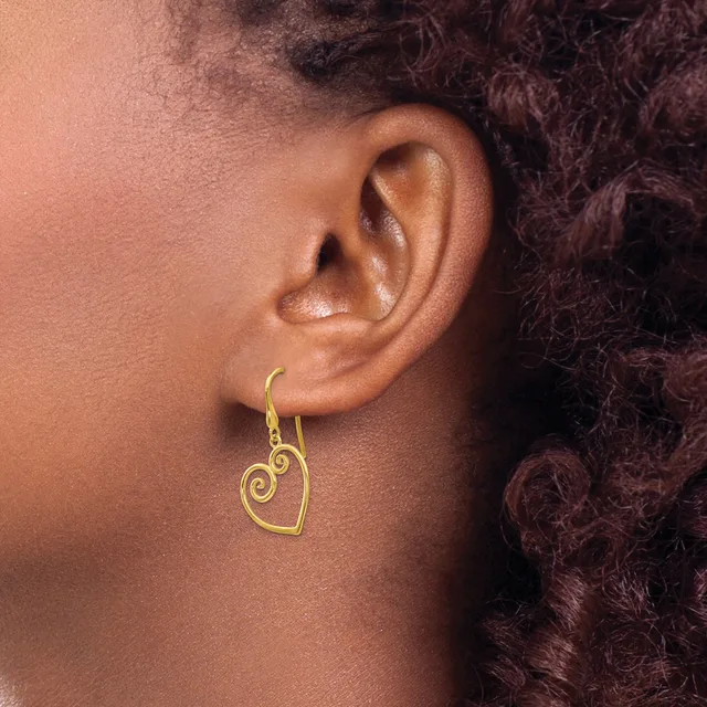 A gold-toned heart-shaped earring with a decorative spiral design hangs from a hook on a person's earlobe.