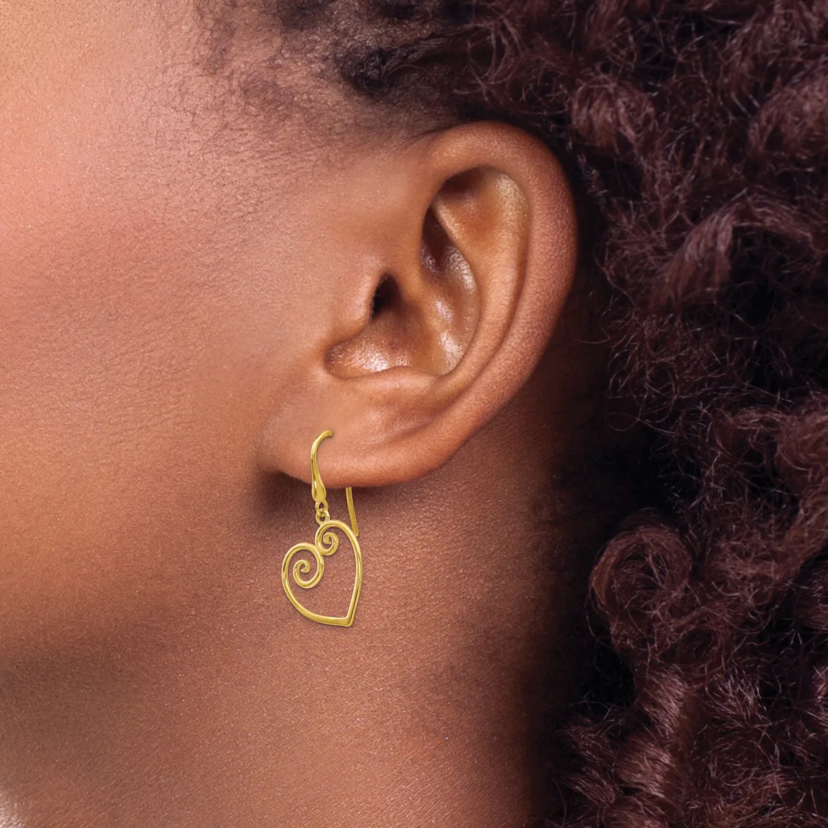 A gold-toned heart-shaped earring with a decorative spiral design hangs from a hook on a person's earlobe.