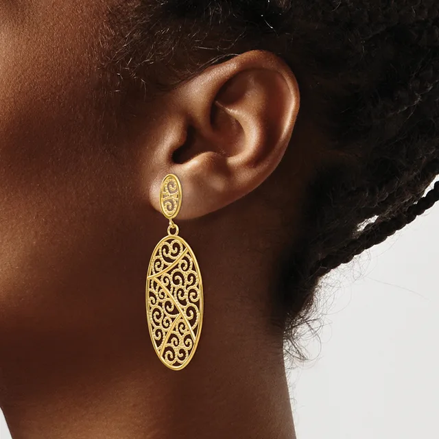 A woman wearing a gold filigree drop earring with an oval stud top and larger ornate oval pendant with scrollwork cutout design.