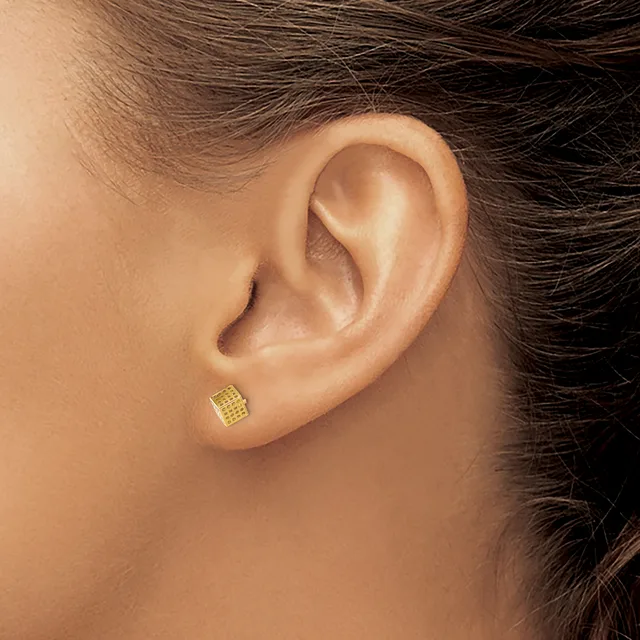 A small square gold stud earring with a textured grid pattern worn in the earlobe of a person with brown hair.