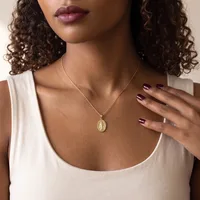 Woman wearing a white sleeveless tank top and a gold pendant necklace with an oval charm, displaying burgundy nail polish on her manicured nails.