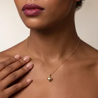 A woman with deep brown skin and burgundy lips wears a gold chain necklace with a small puffy heart pendant, resting her hand on her chest.