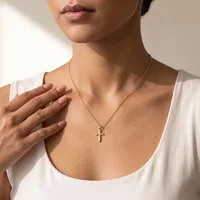 A woman wearing a white tank top displays a delicate gold chain necklace with a small cross pendant resting at her chest.