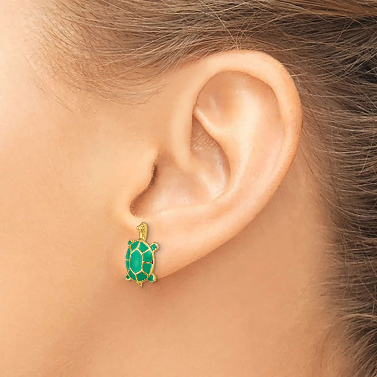 A green enamel turtle charm earring with gold-tone metal details hangs from a pierced earlobe.