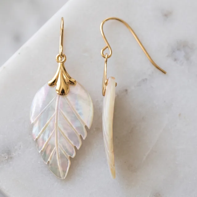 Two mother-of-pearl leaf-shaped drop earrings with carved vein detail and gold-tone hook wires and cap fittings, displayed on white marble, one face-on and one sideways.