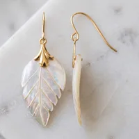 Two mother-of-pearl leaf-shaped drop earrings with carved vein detail and gold-tone hook wires and cap fittings, displayed on white marble, one face-on and one sideways.