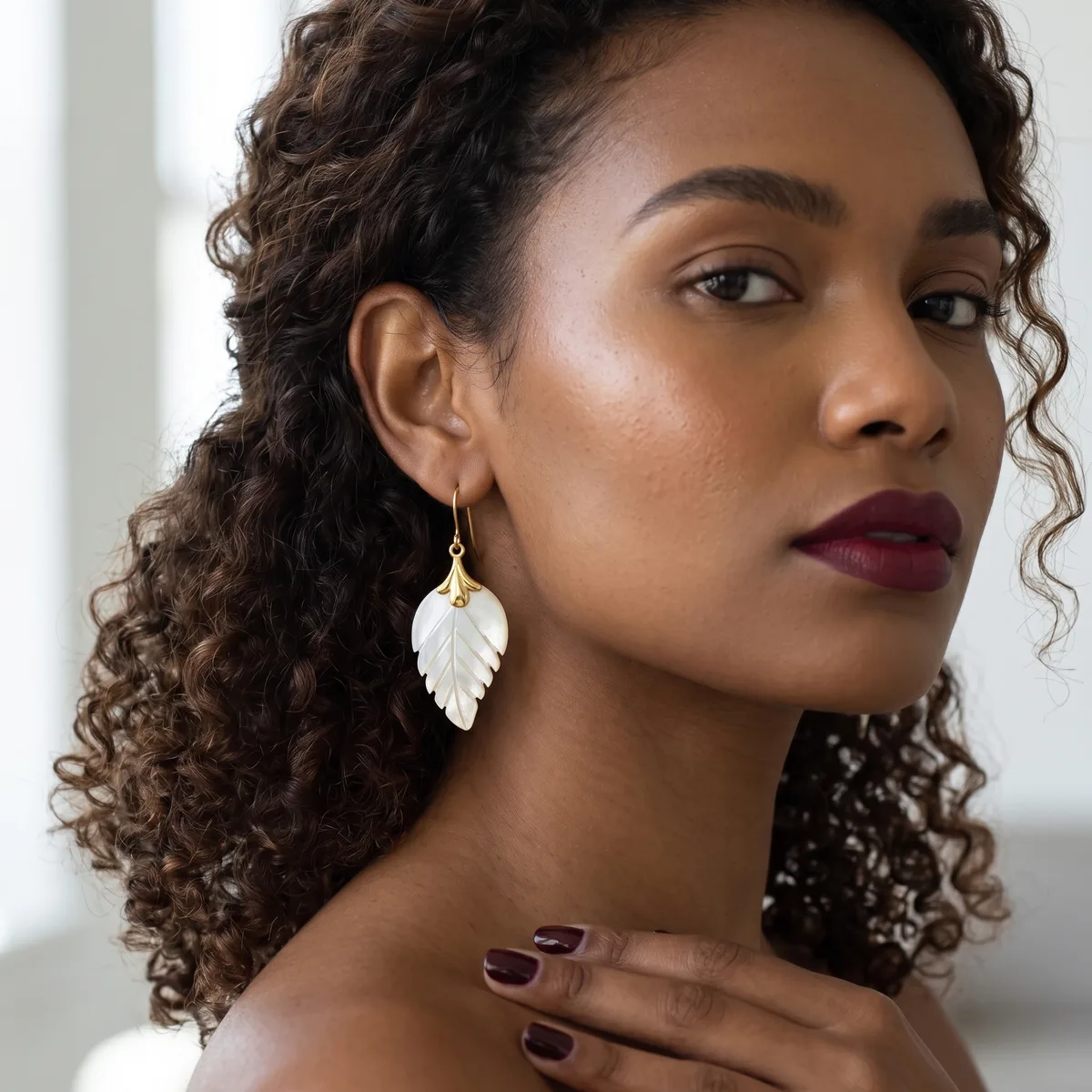 A woman with curly hair wearing a gold hook earring with a carved white mother-of-pearl leaf pendant, shown with dark red lipstick and matching nail polish.