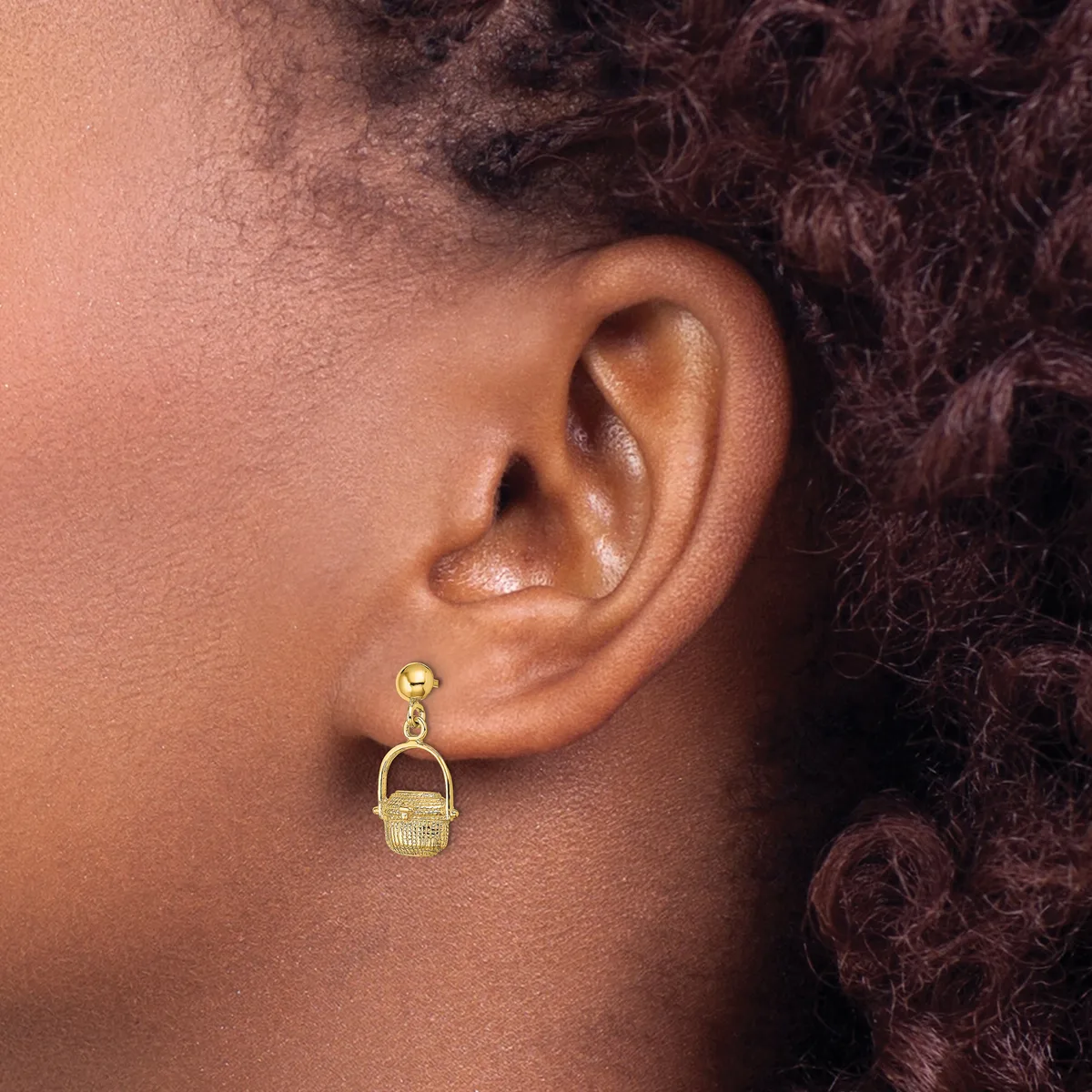 A person wearing a gold-toned drop earring with a small ball post and a textured rectangular pendant hanging below, shown from the side view of their ear.