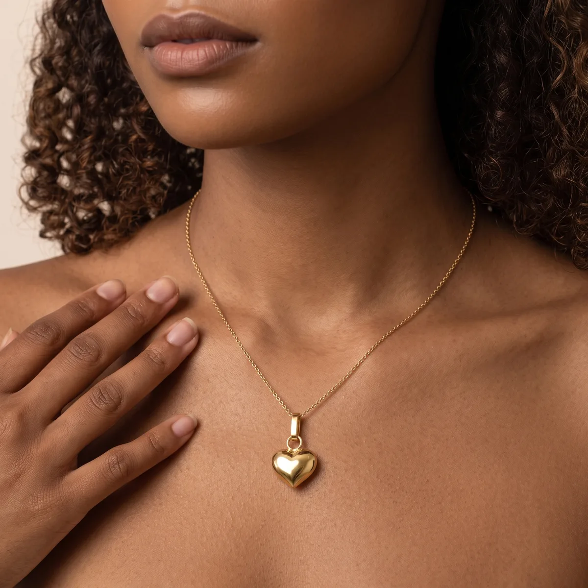 A woman with curly brown hair wears a gold heart pendant necklace and touches her chest with her hand.