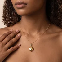 A woman with curly brown hair wears a gold heart pendant necklace and touches her chest with her hand.