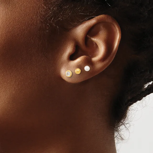 Close-up side view of an ear with three small stud earrings in varying finishes: a sparkly stone, a gold disc, and a white pearl.