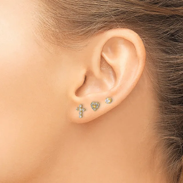 A person wearing three small stud earrings in the lower lobe: a cross with gemstones, a heart with yellow stones, and a round clear stone.