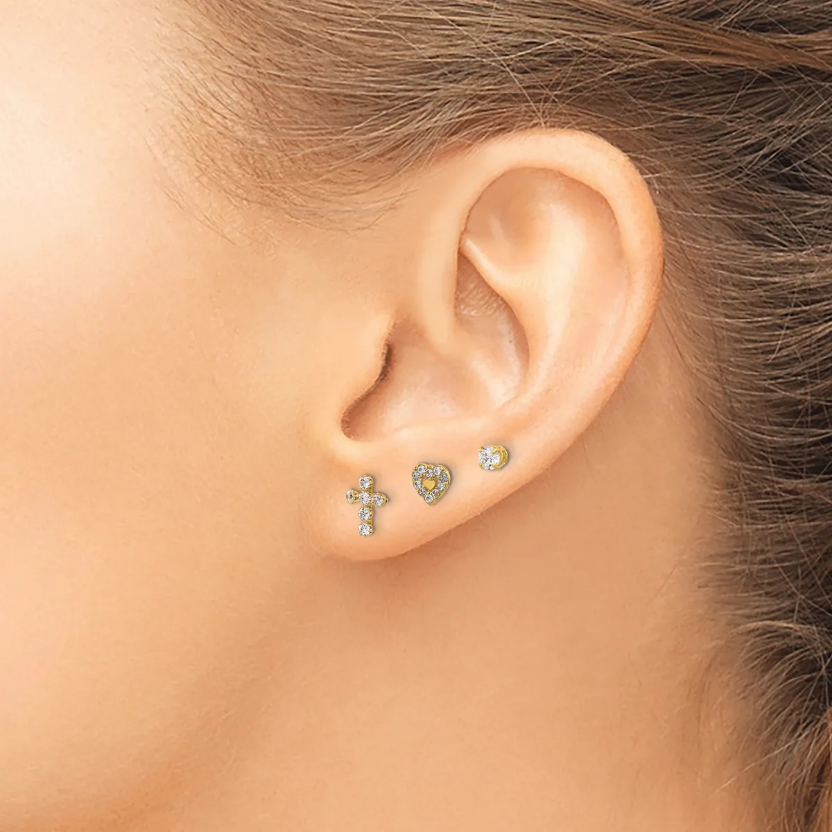 A person wearing three small stud earrings in the lower lobe: a cross with gemstones, a heart with yellow stones, and a round clear stone.