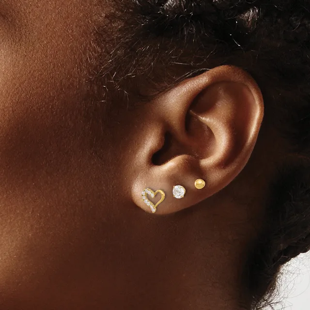 Close-up of a person's ear wearing three small earrings: a gold heart outline, a diamond stud, and a gold disc stud.