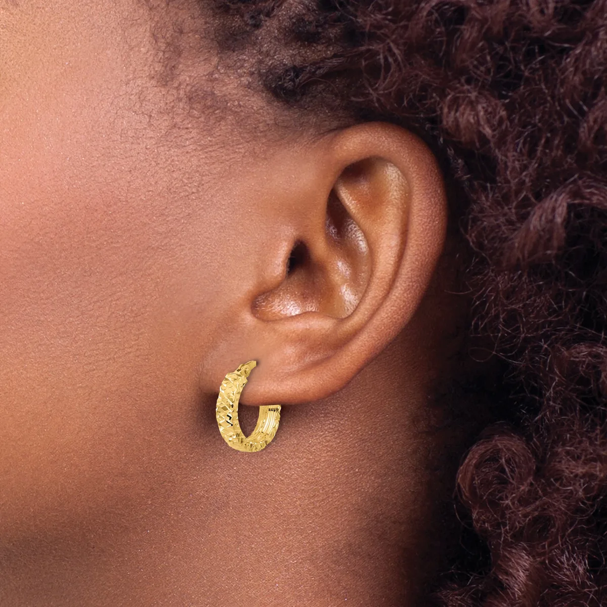 A gold textured hoop earring worn on the earlobe of a person with medium brown skin tone and dark curly hair.