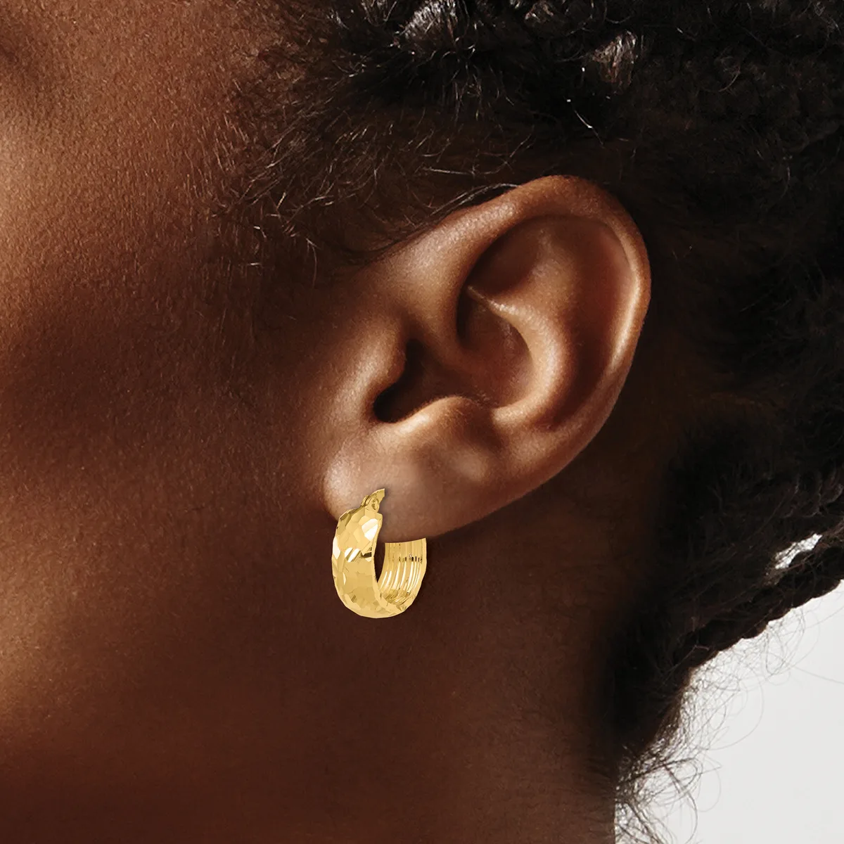 A person wearing a pair of textured gold hoop earrings with a brushed finish, shown from the side profile view against their ear.
