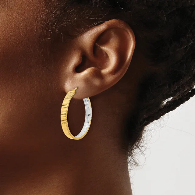 A close-up view of a person's ear wearing two-tone hoop earrings with a textured gold finish on one half and a smooth silver finish on the other half.