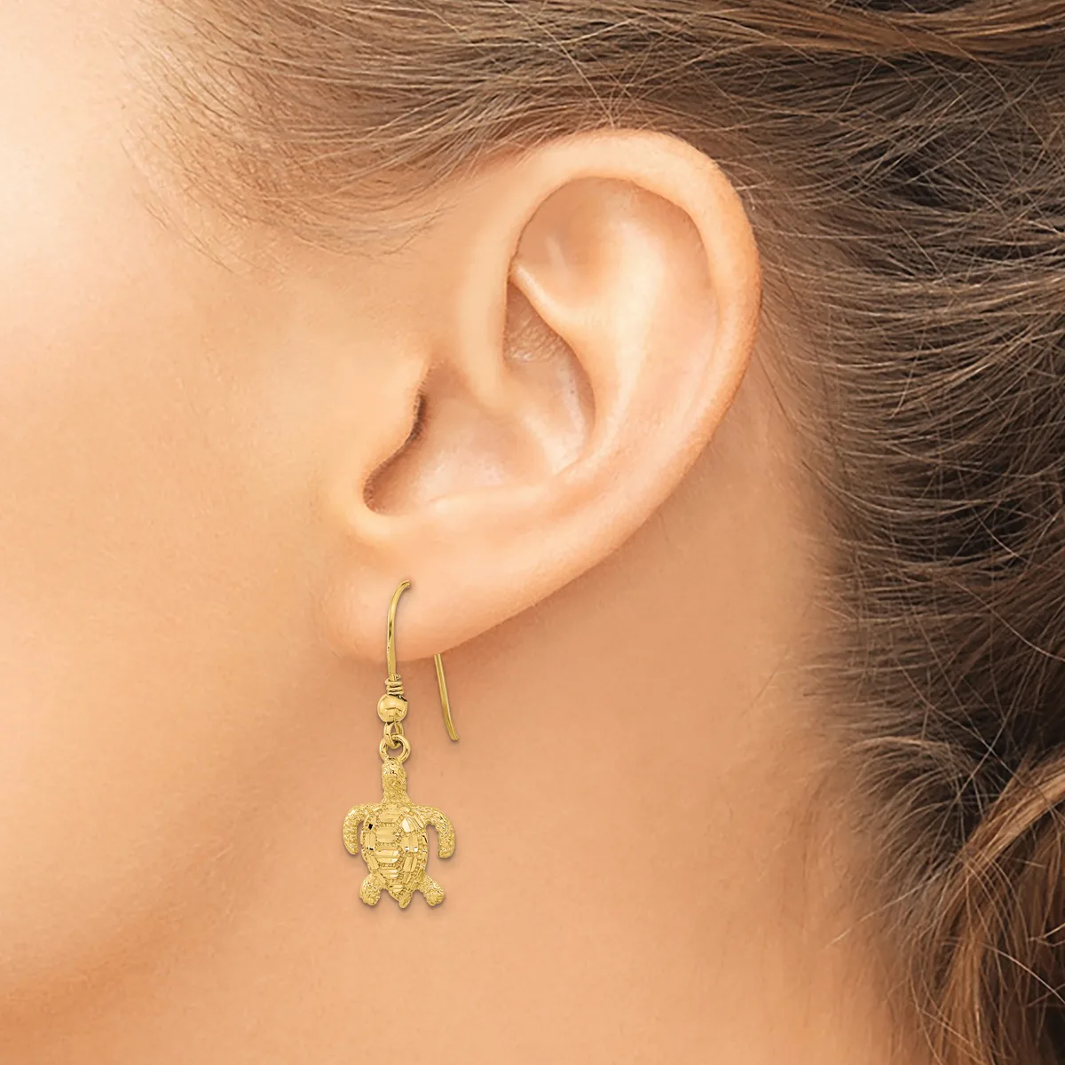 A woman wearing a gold turtle-shaped dangle earring on a hook, shown from the side view of her ear and neck.