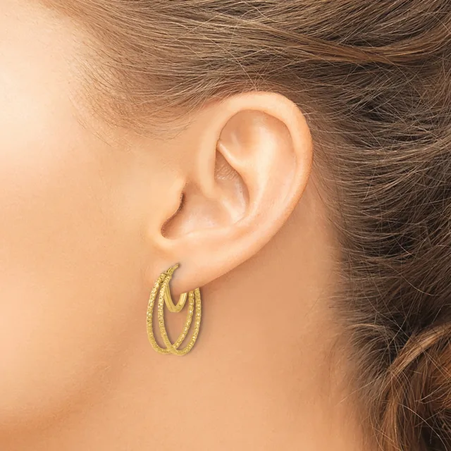 A woman wearing double oval hoop earrings with a textured finish in gold tone metal, shown from the side profile of her face and ear.