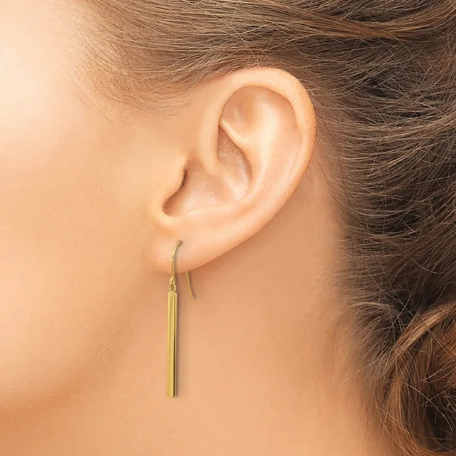 A person wearing a long, thin gold-colored drop earring on a fish hook ear wire, shown from the side view of their ear.