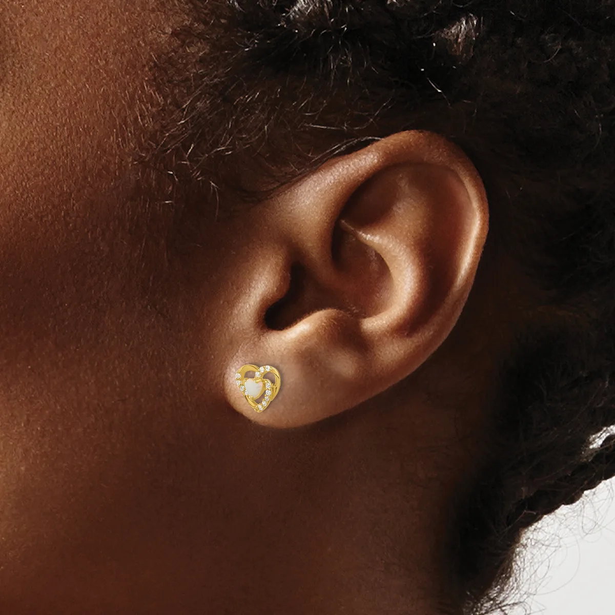 A close-up of a person's ear wearing a small gold heart-shaped stud earring with a white pearl or opal center.