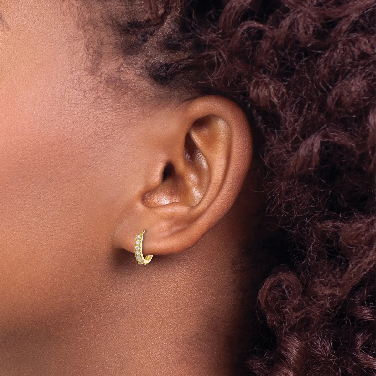 A person wearing a small gold hoop earring with diamond or crystal embellishments in their earlobe, shown in profile against dark curly hair.