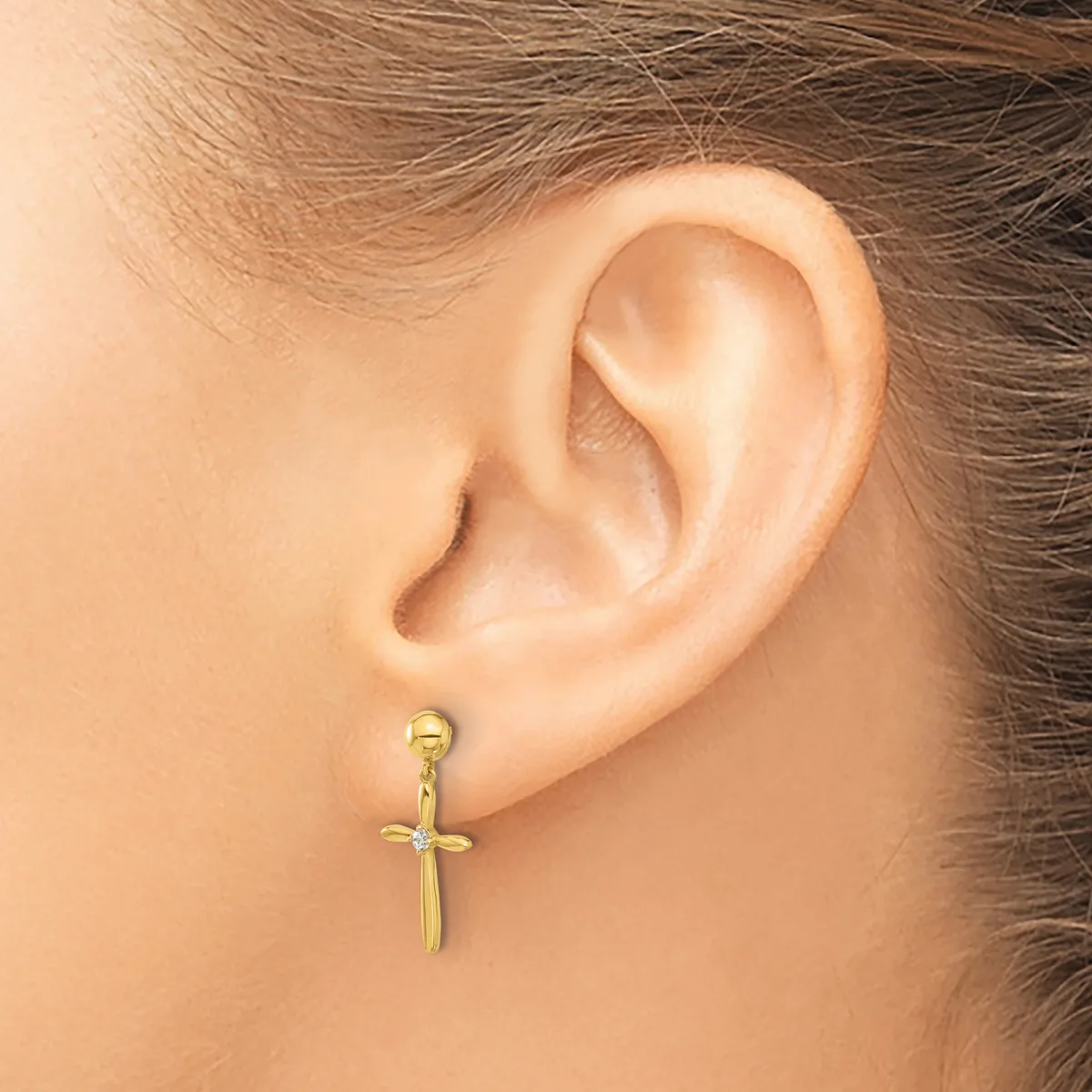 A gold cross-shaped earring hangs from a pierced earlobe on a person with brown hair.