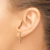 A gold cross-shaped earring hangs from a pierced earlobe on a person with brown hair.