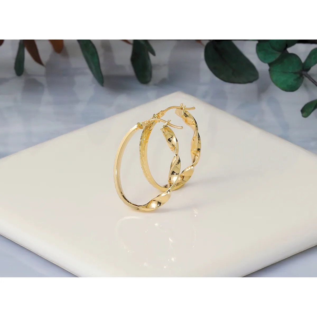 A pair of gold hoop earrings with a twisted or textured surface pattern, displayed on a white ceramic dish with green foliage in the background.