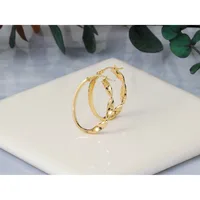 A pair of gold hoop earrings with a twisted or textured surface pattern, displayed on a white ceramic dish with green foliage in the background.
