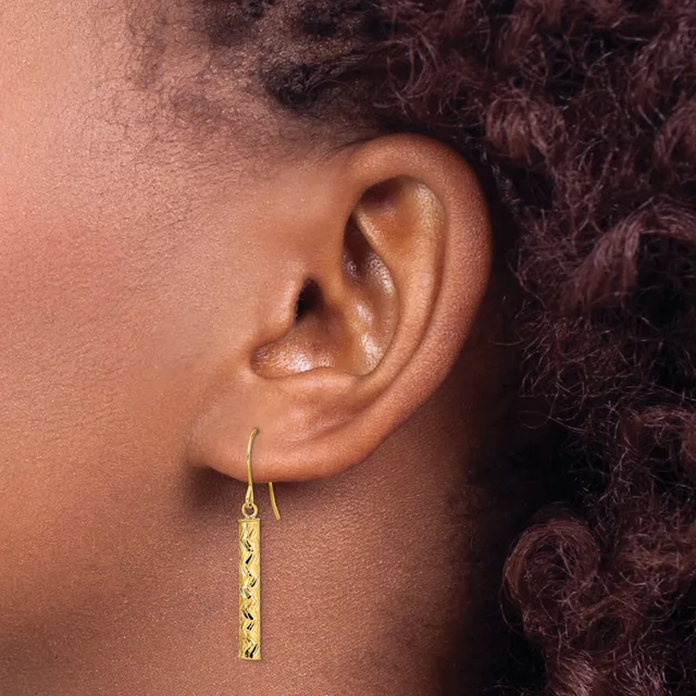A person wearing gold drop earrings with a textured rectangular pendant hangs from a curved hook through their earlobe.