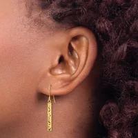 A person wearing gold drop earrings with a textured rectangular pendant hangs from a curved hook through their earlobe.