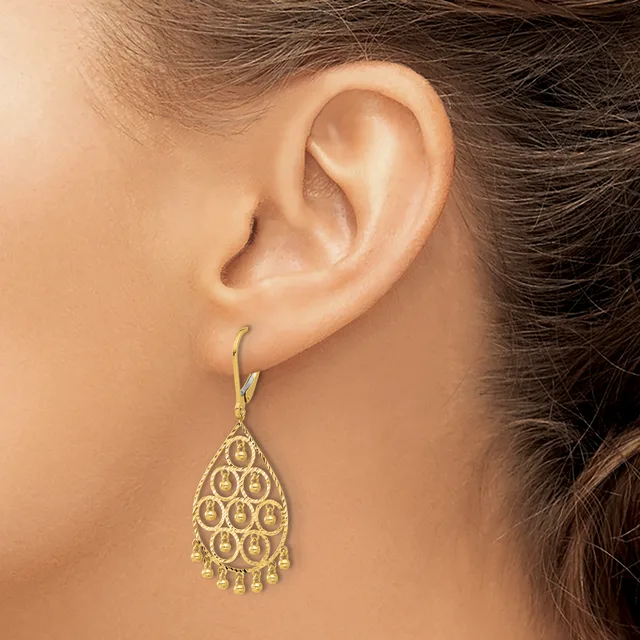 A woman wearing a gold teardrop-shaped dangle earring with a filigree pattern and small beads hanging from the bottom.