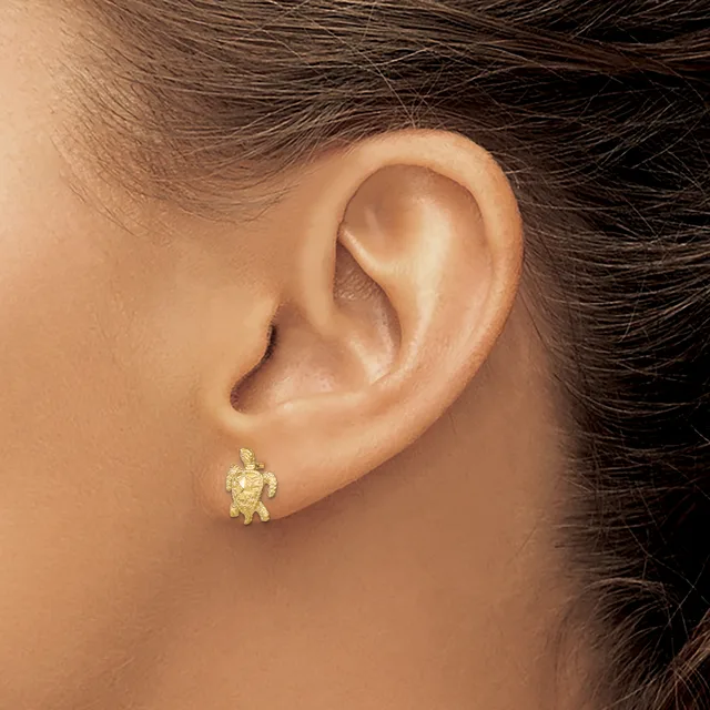 A small gold turtle-shaped stud earring worn on a person's earlobe, shown in close-up against their ear and shoulder.