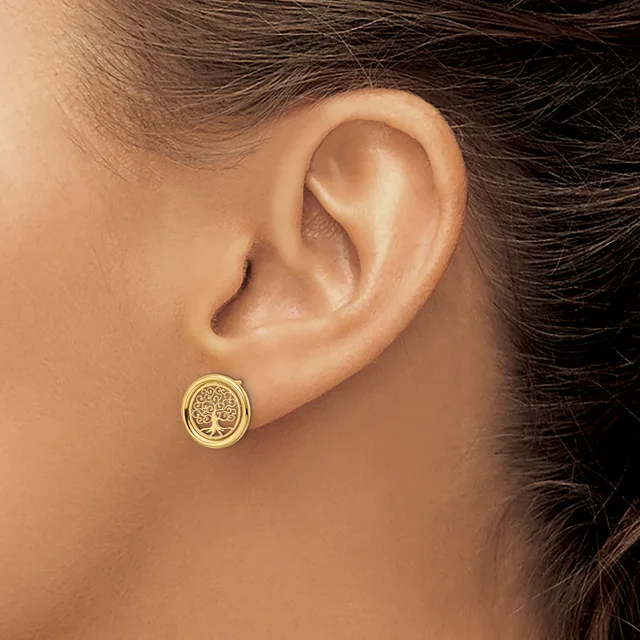 A person wearing a small gold circular stud earring with a tree design in the center, shown from the side view of their ear.
