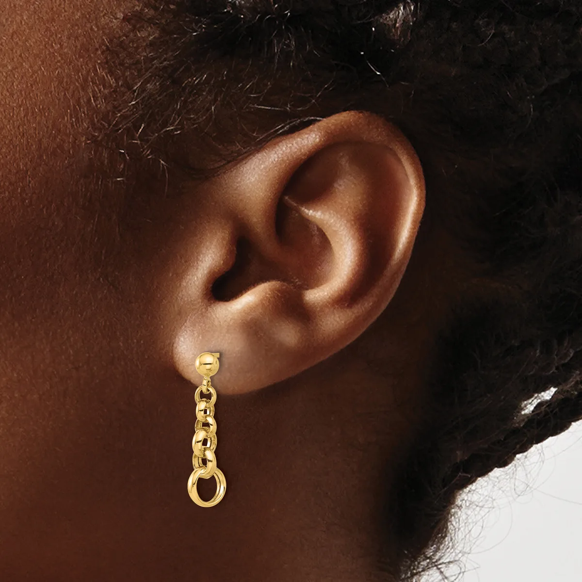 A gold chain drop earring with a small loop at the top, worn on the earlobe of a person with brown skin and dark hair.