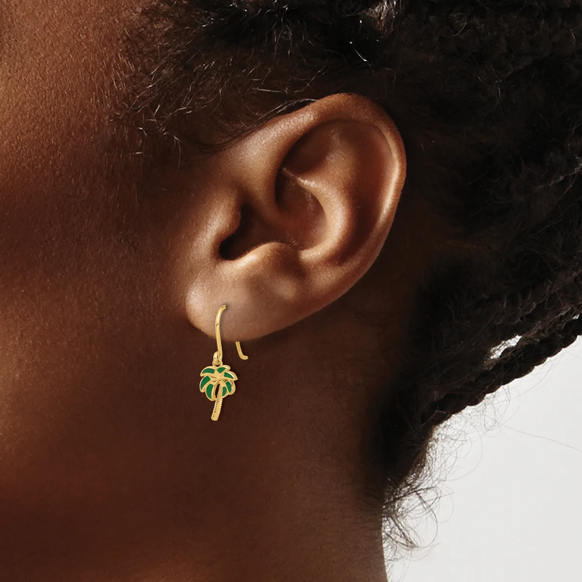 A person wearing a gold drop earring with a green and red enamel cross design hanging from a curved wire hook.
