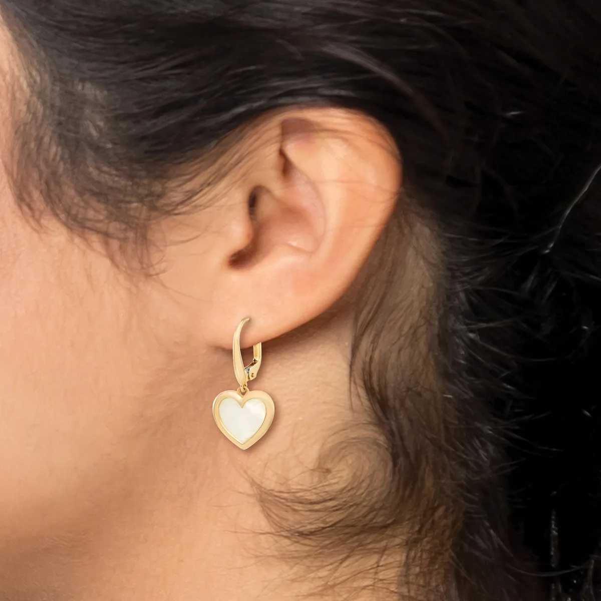 A person wears a gold hoop earring with a dangling heart-shaped pendant in white stone or enamel on their earlobe.
