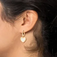 A person wears a gold hoop earring with a dangling heart-shaped pendant in white stone or enamel on their earlobe.