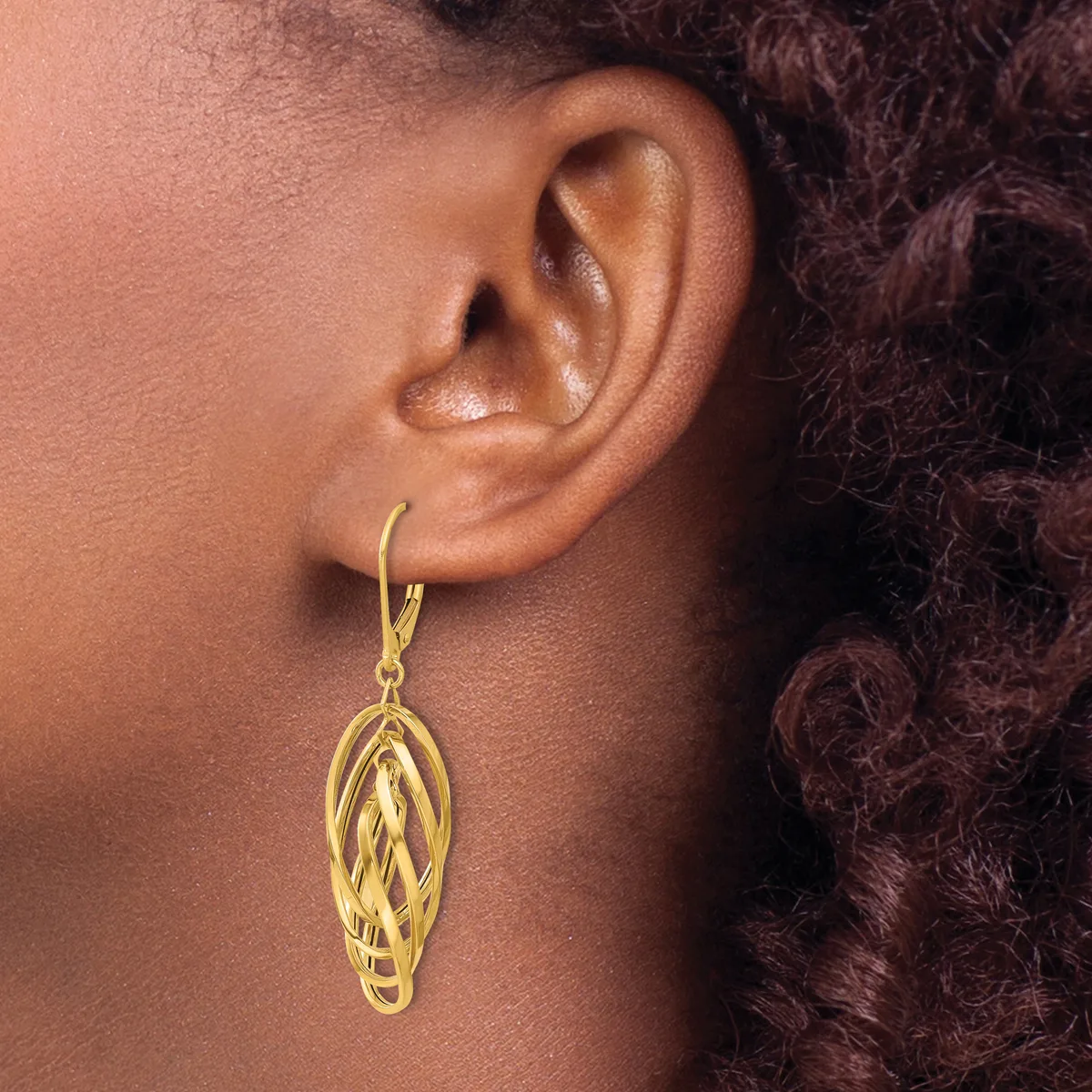 A person wearing a gold-colored dangle earring with an elongated oval design featuring twisted or woven detailing, shown from the side view against their neck and shoulder.