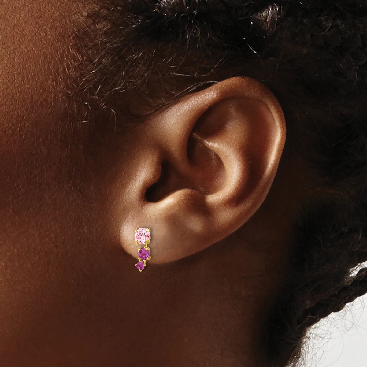 A person wearing a gold drop earring with purple oval stones arranged vertically, shown from the side profile view of their ear.